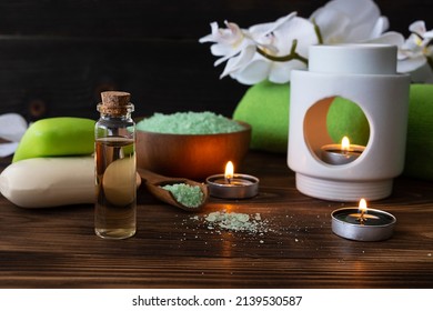 Body oil, tea candles and bath soap on a dark brown wooden table. Green bath towel and orchid sprig in the background