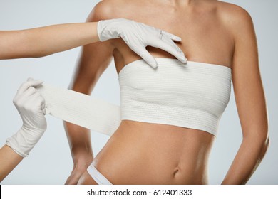 Body Plastic Surgery. Slim Young Female Body In White Panties And Breast Bandages On White Background. Closeup Of Doctor Hands In Gloves Wrapping Female Patient's Breast In Bandage. High Resolution