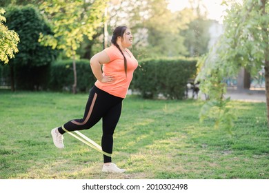 Body Positive. Fat Burning. Park Exercising. Band Workout. Cheerful Diligent Overweight Obese Woman In Activewear Training Legs In Green Spring Park.