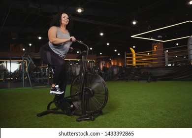 Body Positive, Feminine Confidence Concept. Full Length Shot Of Overweight Beautiful Happy Woman Cycling At The Gym On Air Bike. Cheerful Plus Size Woman Doing Cardio Workout On Air Cycle, Copy Space