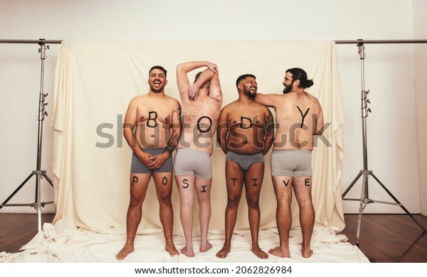 Body positive men wearing underwear in a studio.\
Four happy men standing with the letters that read’ ’BODY\
POSITIVITY” written on their bodies. Self-confident young men\
embracing their natural\
bodies.