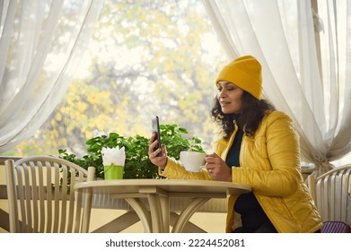 Body positive multi-ethnic beautiful woman in yellow hat and jacket, sitting at table with hot aroma coffee, talking on smartphone by video link, having an online meeting with friends from a cozy cafe - Shutterstock ID 2224452081