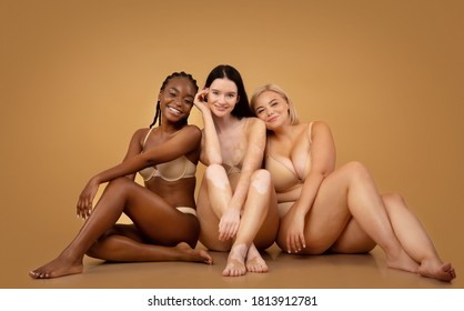 Body Positive. Three attractive multicultural women in lingerie with different skin and body type sitting on floor, posing together in studio