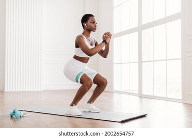 Body Shaping And Glutes Workout. Determined Fit Black Woman In White Sportswear Exercising With Resistance Loop Band Doing Squats Standing On Yoga Mat, Training Butt And Hamstring, Wearing Earbuds