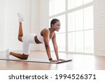 Body Shaping And Glutes Workout. Smiling Fit Black Woman In White Sportswear Doing Donkey Kicks Exercise With Leg Raise On Yoga Mat, Training Butt And Hamstring, Blurred Background. Fitness Concept