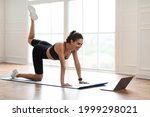 Body Shaping And Glutes Workout. Smiling Fit Woman In Black Sportswear Doing Donkey Kicks Exercise With Leg Raise On Yoga Mat, Training Butt And Hamstring, Looking At Laptop Screen On Floor