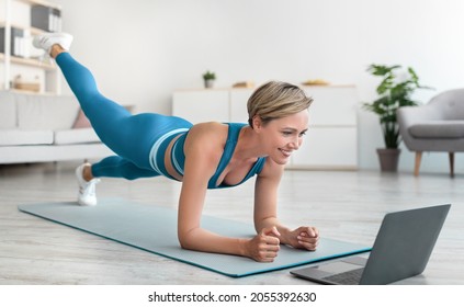 Body Shaping And Glutes Workout. Smiling Fit Woman In Blue Sportswear Standing In Plank Pose With Leg Lift, Exercising On Yoga Mat, Training Butt And Hamstring, Watching Video Tutorial On Laptop