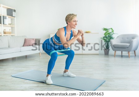 Body Shaping And Glutes Workout. Smiling Fit Woman In Blue Sportswear Exercising With Resistance Loop Band Doing Squats Standing On Yoga Mat, Training Butt And Hamstring In Living Room At Home