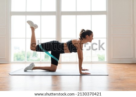 Body Shaping And Glutes Workout. Smiling Fit Woman Doing Donkey Kicks Exercise With Leg Raise On Yoga Mat Using Resistance Band, Training Butt Muscles And Hamstring. Profile Side View. Fitness Concept