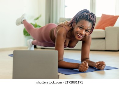 Body Shaping Workout. Fit Black Woman Standing In Plank Pose With Leg Lift, Exercising On Yoga Mat And Watching Video Tutorial On Laptop, Smiling At Camera