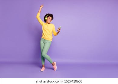 body size photo of curly wavy trendy sweet cute charming carefree girl dancing and singing while reading song text on her telephone and wearing headphones isolated violet pastel color background