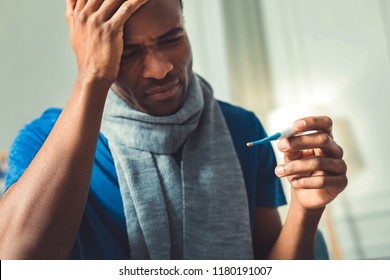 Body Temperature. Low Angle Of Concerned Afro American Man Looking At Thermometer And Touching Head