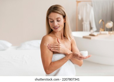 Body treatments. Happy middle aged woman applying moisturising cream on shoulder, sitting wrapped in towel and making beauty care routine at home, copy space - Powered by Shutterstock