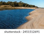 A body of water with a sandy beach and trees in the background. The water is calm and blue