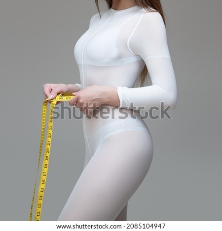 body of a woman in a white suit for LPG massage holds a centimeter in her hands. Anti-cellulite body care concept. Gray background
