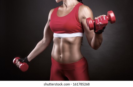 Body of a young fit woman lifting dumbbells in a red sportswear on a dark background
