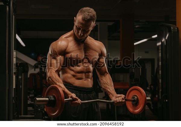 A bodybuilder with a beard is doing bicep curls\
with an EZ-barbell in a gym. A photo of a torso of a naked muscular\
man during a workout.