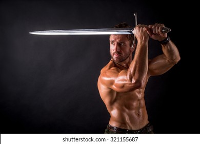 Bodybuilder man posing with a sword isolated on black background. Serious shirtless man demonstrating his mascular body.