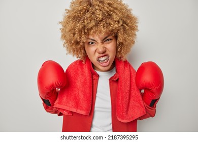 Bodybuilding And Sport Concept. Angry Curly Female Boxer Wears Boxing Gloves Puts On Mouthguard Ready To Fight And Become Winner Of Championship Going To Give Punch Isolated Over Grey Background