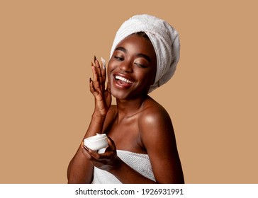 Bodycare Concept. Positive African American Young Woman Applying Beauty Product On Her Face. Pretty Smiling Black Lady Wrapped In Towel Using Cream After Shower, Isolated On Studio Background.