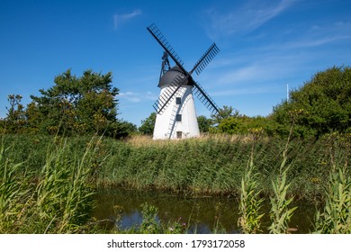 Bogense, Denmark-August 2020; Typical White Windmill For The Funen Area Close To Bogense With In The Foreground The Water Of A Canal With Beds Of Reed Against A Blue Sky