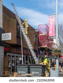 Bognor Regis, Sussex, UK - 18 Mar 2021: A Firefighter Climbs A Ladder Towards The Roof Of A Shop, Colleagues Stand And Support The Base. Scaffold With Red Safety Net Against A Blue Sky - Copy Space. 