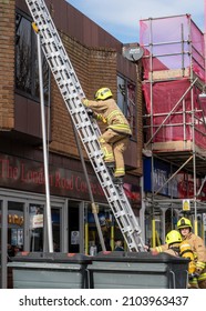 Bognor Regis, Sussex, UK - 18 Mar 2021: A Firefighter Climbs A High Ladder Against A Red Brick Shop Building, Colleagues Stand And Support The Base. Behind Scaffold Wrapped In Red Safety Netting.