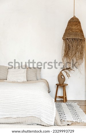 bohemian interior in bedroom. comfort bed with cushions and blanket, ceramic vase with dry plants on wooden table and decorative wicker lampshade against white copy space wall