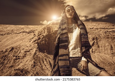 Boho Chic Fashion. A Beautiful Woman In Elegant Boho Style Clothes And Jewelry With An Ethnic Headscarf On Her Head Stands Against The Backdrop Of Sand Dunes And Bright Sun In The Sky.