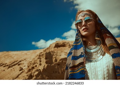 Boho Chic Fashion, Summer Style. A Beautiful Woman In Elegant Boho Style Jewelry With An Ethnic Headscarf On Her Head Stands Against The Backdrop Of Sand Dunes And Blue Sky. Copy Space.