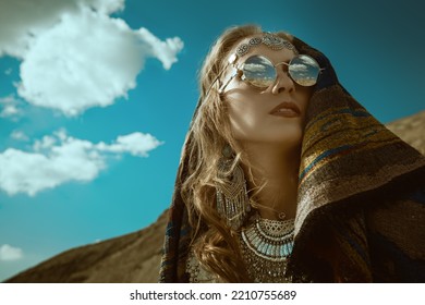 Boho Chic Fashion, Summer Style. A Beautiful Woman In Elegant Boho Style Jewelry With An Ethnic Headscarf On Her Head Stands Against The Backdrop Of Sand Dunes And Blue Sky. Copy Space.