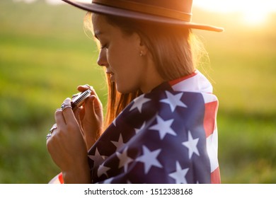 Boho Chic Woman In Hat With American Flag Playes On Harmonica Outdoors At Sunset. Jewelry Indie Girl With Hippie Style And Boho Fashion. Travel To America