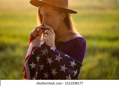 Boho Chic Woman In Hat With American Flag Playes On Harmonica Outdoors At Sunset. Jewelry Indie Girl With Hippie Style And Boho Fashion. Travel To America