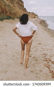 Boho Girl In White Shirt Walking On Sunny Beach, Back View. Carefree Stylish Woman In Swimsuit And Shirt Relaxing On Seashore. Summer Vacation. Lifestyle Authentic Image