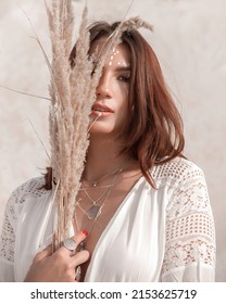 Boho Style Portrait. Young Brunette Girl In White Boho Dress Stands Casual With Beige Dried Flower Near Face On A White Sand Career Background And Smiles Cute. Authentic Lifestyle Concept, Free Space