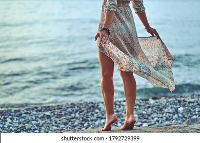 Boho woman with long legs and waving dress standing on a stone by the seashore at sunset 