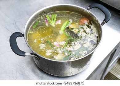 Boiled down broth cooked from vegetable scraps and poultry trimmings with protein foam and fat eyes in a large stainless steel pot on a professional kitchen counter, selected focus