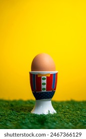 A boiled egg in an egg cup, on artificial grass and in front of a yellow background.