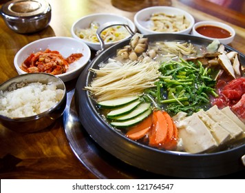 Boiled Korean food consisting of meat, ginseng, mushrooms and vegetables. There are rice, dipping sauce and some pickled bean sprouts on a wooden table.