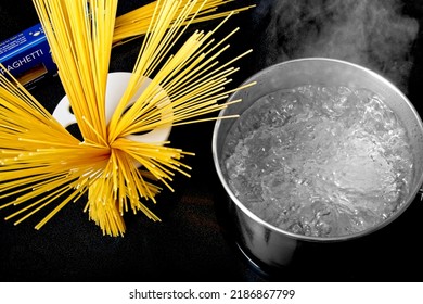 Boiling Pot Of Spaghetti Water On Stovetop With Steam Emanating From Hot Water. Including Spiraled Spaghetti Noodles In A Portion Measuring Ring And Pasta Packaging In Background.