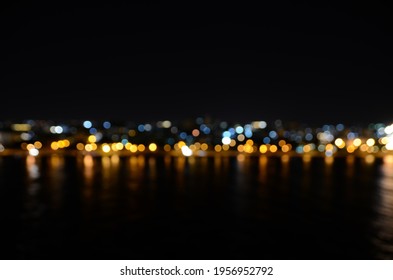 Bokeh Of Night Coastline Scene. Blur Effect From The Water And City Lights In The Background Reflecting In The Water. Defocused City Skyline In Front Of The Ocean.
