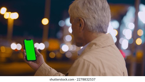 Bokeh shot of aged man holding smartphone with blank green screen outdoors. Portrait of retired male using mobile phone stranding on city street at night - Powered by Shutterstock