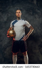 Bold and handsome soccer player posing for a photoshot in a dark studio, wearing professional sportswear, and holding a golden soccer ball, full height