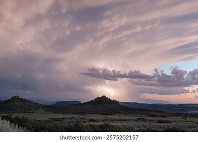 A bolt of lightning drops from clouds tinged with the pink of dusk and arcs above a rocky knoll on Kolob Mt. in Southern Utah, USA during a summer monsoon storm while a touch of clear sky peek through