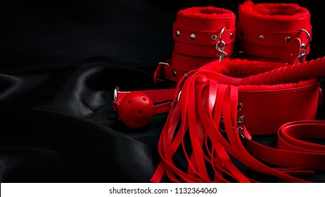 Bondage, Kinky Adult Sex Games, Kink And BDSM Lifestyle Concept With A Pair Of Red Leather Handcuffs, Flogger, Ball Gag And A Coller With A Leash Attached On Black Silk With Copy Space