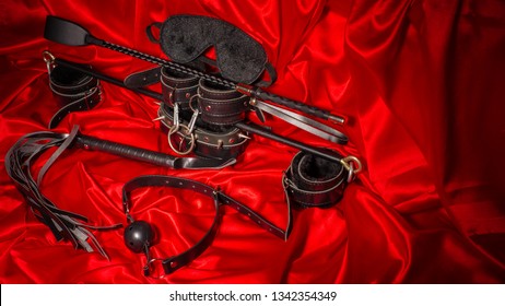 Bondage, Kinky Adult Sex Games, Kink And BDSM Lifestyle Concept With A Pair Of Leather Handcuffs, Flogger, Ball Gag And A Coller With A Leash Attached On Red Silk With Copy Space