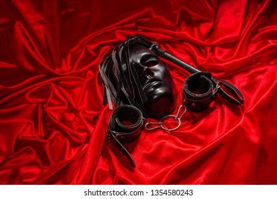 Bondage, kinky adult sex games, kink and BDSM lifestyle concept with a mask, pair of leather handcuffs, flogger, ball gag and a coller with a leash attached on red silk with copy space