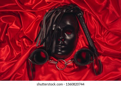 Bondage, Kinky Adult Sex Games, Kink And BDSM Lifestyle Concept With A Mask, Pair Of Leather Handcuffs, Flogger, Ball Gag And A Coller With A Leash Attached On Red Silk With Copy Space