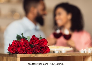 Bonding Concept. Bouquet Of Flowers Lying On Table, Selective Focus On Red Roses. Happy Black Man And Woman Spending Time Together At Home Or Cafe, Having Dinner, Drinking Wine In Blurred Background