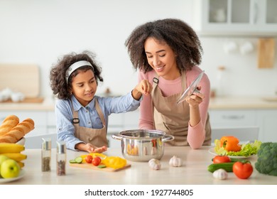 Bonding Concept. Cheerful african american mother helping her daughter to cook, teaching little girl how to prepare healthy vegetable soup, salting water in pot, cooking together at home. - Shutterstock ID 1927744805